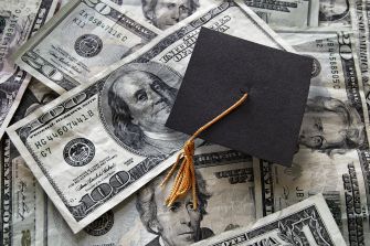 Graduation cap on top of a pile of one-hundred dollar bills representing student loans.
