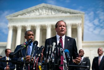 Texas officials speaking at a podium with microphones in front of a large government building.