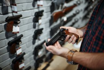 A person examining a handgun in a firearm store, with various guns displayed on a wall behind them.