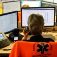 A person speaking on the phone while sitting in an emergency call center surrounded by multiple computer monitors. A person speaking on the phone while sitting in an emergency call center surrounded by multiple computer monitors.