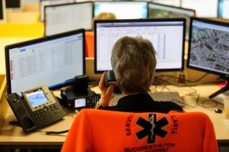 A person speaking on the phone while sitting in an emergency call center surrounded by multiple computer monitors.