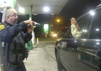 A police officer points a gun at a man with his hands raised inside a car during a traffic stop at a gas station.
