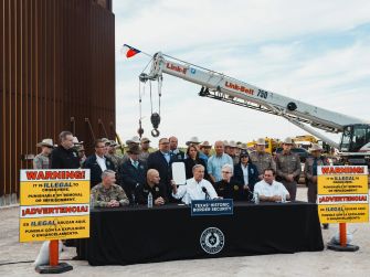 Texas Governor Greg Abbott signing a bill related to border security, surrounded by law enforcement and supporters, with warning signs about illegal immigration in the foreground.