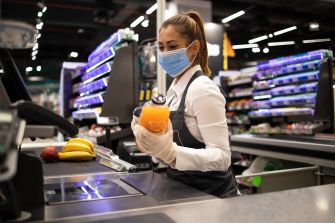 A cashier wearing a mask and gloves scans groceries at a checkout counter in a store.