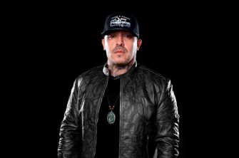 Image of a man wearing a black leather jacket and a cap, against a dark background.