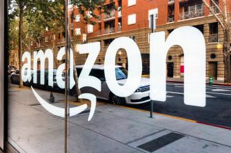 Amazon logo displayed on a storefront window with a city street view in the background.