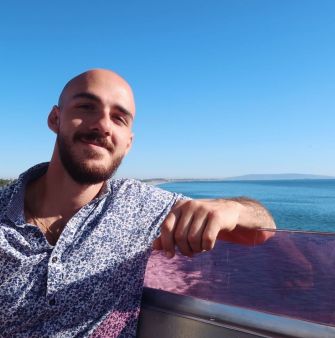 A young man sitting by the water, smiling and wearing a patterned shirt.