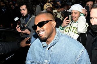 Kanye West surrounded by fans and photographers, smiling while wearing sunglasses and a denim jacket.