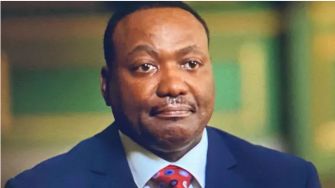 A man in a suit with a concerned expression on his face, seated in a setting with muted colors.