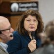 A woman speaking into a microphone at a library meeting, with a blurred audience in the foreground. A woman speaking into a microphone at a library meeting, with a blurred audience in the foreground.