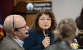 A woman speaking into a microphone at a library meeting, with a blurred audience in the foreground.