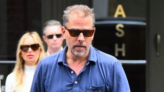 Hunter Biden walking outside wearing sunglasses, accompanied by a woman in the background.