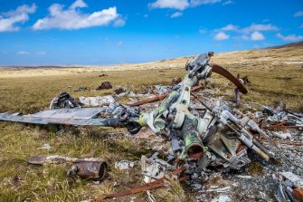 Wreckage of a helicopter crash site in a grassy landscape, with debris scattered under a blue sky.