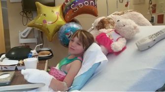 A young girl recovering in a hospital bed surrounded by stuffed animals and get well balloons.
