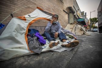 A person sitting in front of a tent in an urban environment, surrounded by belongings.