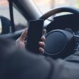 A driver holding a smartphone while gripping the steering wheel of a car. A driver holding a smartphone while gripping the steering wheel of a car.
