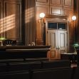 A courtroom interior featuring wooden paneling, rows of empty seats, and a view of the judge's bench with light streaming in through the windows. A courtroom interior featuring wooden paneling, rows of empty seats, and a view of the judge's bench with light streaming in through the windows.