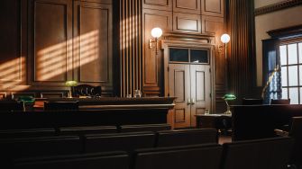 A courtroom interior featuring wooden paneling, rows of empty seats, and a view of the judge's bench with light streaming in through the windows.