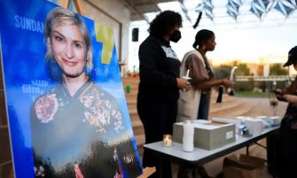 A woman stands next to a photograph of Halyna Hutchins at an event, while others are engaged in activities at a table.