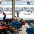 A group of migrants resting on cots surrounded by blankets inside a busy terminal, with people walking past outside. A group of migrants resting on cots surrounded by blankets inside a busy terminal, with people walking past outside.
