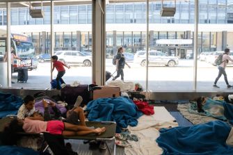 A group of migrants resting on cots surrounded by blankets inside a busy terminal, with people walking past outside.
