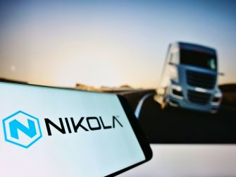 Image of a smartphone displaying the Nikola Motors logo, with a blurred image of a truck on a road in the background.