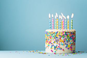 A colorful birthday cake with lit candles and vibrant sprinkles against a light blue background.