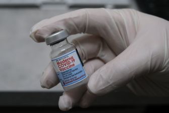 A gloved hand holding a vial of the Moderna COVID-19 vaccine.