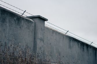 A gray prison wall with barbed wire against a cloudy sky.