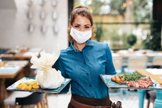 A waitress wearing a mask holds two plates of food in a restaurant setting.