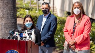 A press conference setting with three individuals discussing California's guaranteed income program, with microphones and a podium in view.