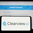 Clearview AI facial recognition software logo on the glowing screen and blurred faces from social media on the background.