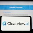 A hand holding a smartphone displaying the Clearview AI logo in front of a screen showing the company's website. A hand holding a smartphone displaying the Clearview AI logo in front of a screen showing the company's website.