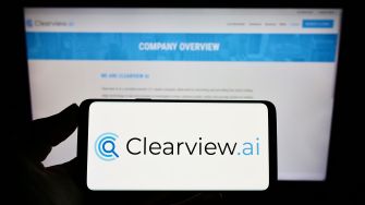 A hand holding a smartphone displaying the Clearview AI logo in front of a screen showing the company's website.