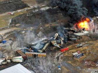 Aerial view of a train derailment site in East Palestine, Ohio, showing smoke and flames from burning tank cars.
