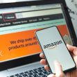 A person holding a smartphone displaying the Amazon logo, with an Amazon shopping website open on a laptop in the background. A person holding a smartphone displaying the Amazon logo, with an Amazon shopping website open on a laptop in the background.