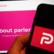 Logo of Parler displayed on a smartphone in front of a computer screen showing information about the platform. Logo of Parler displayed on a smartphone in front of a computer screen showing information about the platform.