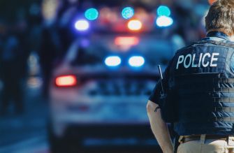 A police officer in a tactical vest stands in front of a police car with flashing lights, suggesting an ongoing law enforcement situation.