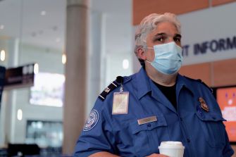 TSA employee wearing a mask while holding a coffee cup at an airport information desk.