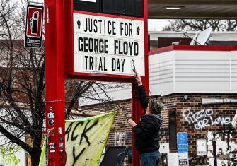 A person adjusts a sign that reads "Justice for George Floyd Trial Day 3" outside a building covered in graffiti.