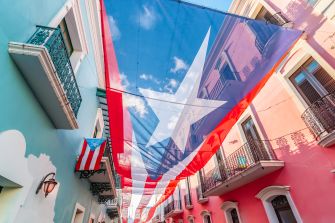 A large Puerto Rican flag hangs between colorful buildings in a street, symbolizing the island's ongoing discussions about statehood and political status.