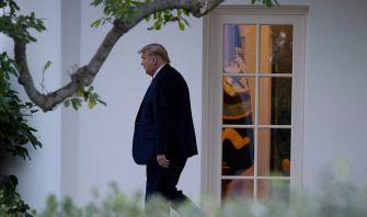 A man in a suit walking outside near a window of a building, with trees and foliage in the foreground.