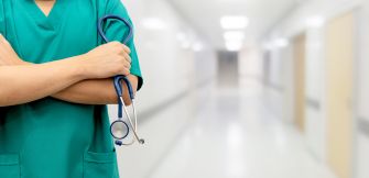 A nurse in scrubs holding a stethoscope, standing in a hospital corridor.