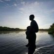 A silhouette of a person carrying a child on their back near a river at sunset, symbolizing the journey of migrants. A silhouette of a person carrying a child on their back near a river at sunset, symbolizing the journey of migrants.
