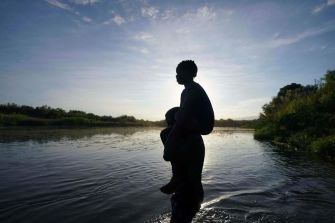 A silhouette of a person carrying a child on their back near a river at sunset, symbolizing the journey of migrants.