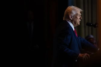 Donald Trump speaking at a podium, partially illuminated against a dark background.