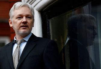 Julian Assange standing near a window, looking out, with his reflection visible.