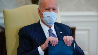 A seated President Biden wearing a mask, holding a binder, during a meeting.