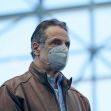 A man wearing a face mask and a brown leather jacket stands in a brightly lit indoor space. A man wearing a face mask and a brown leather jacket stands in a brightly lit indoor space.