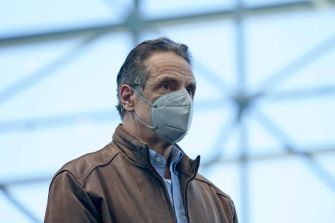 A man wearing a face mask and a brown leather jacket stands in a brightly lit indoor space.